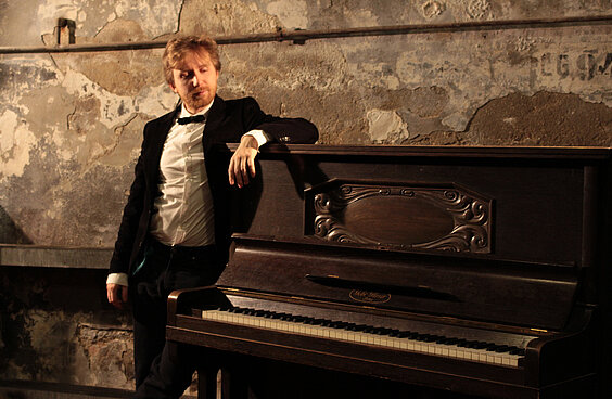 Stummfilmmusiker Richard Siedhoff lehnt im Anzug an einem Klavier vor einer Steinwand