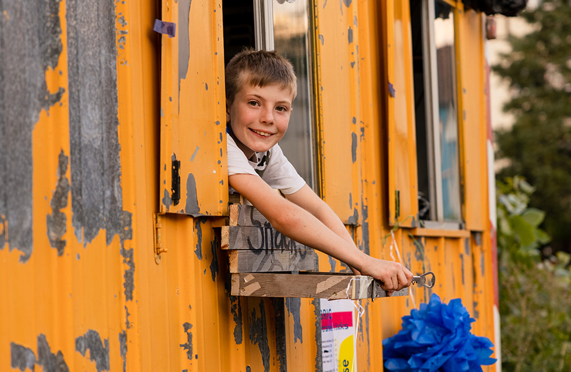 Ein Kind schaut aus dem Fenster von einem gelben Bauwagen, dekoriert mit Pompoms 
