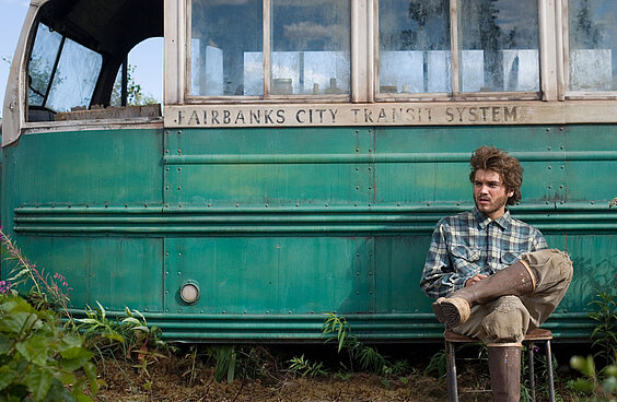 Filmstill Into the Wild Schauspieler Emile Hirsch sitzt als Figur Christopher McCandless mit übereinander geschlagenen Beinen vor einem grünen schmutzigen Van entspannt auf einem Klappstuhl