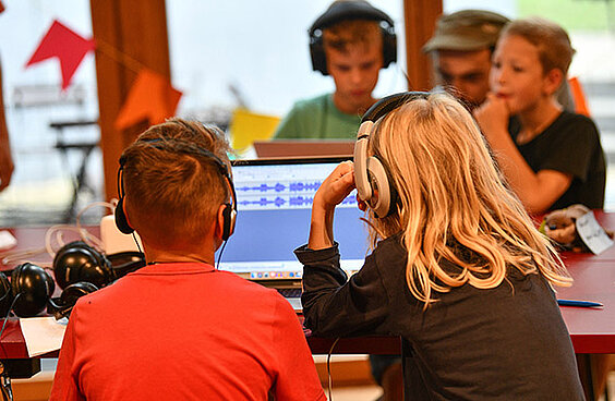 Zwei Kinder mit Kopfhörern schauen auf einen Bildschirm, auf dem ein Audio-Aufnahme Programm zu sehen ist