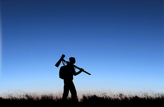 Silouette der Fotografin Kerstin Langenberger mit Kamerastativ über der Schulter vor Abendhimmel