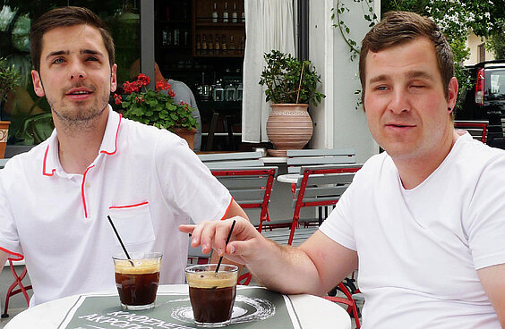 Filmstill Blindflug Die Freunde Yves und Jonas sitzen im Café mit je einem Kaffee im Glas mit Strohhalm 