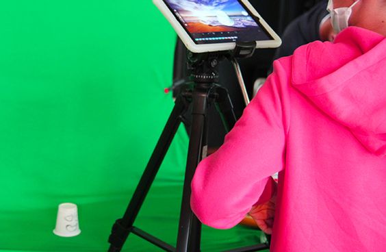 Jemand hält eine Stativ-Halterung mit einem Tablet und filmt einen Becher auf grünem Hintergrund 