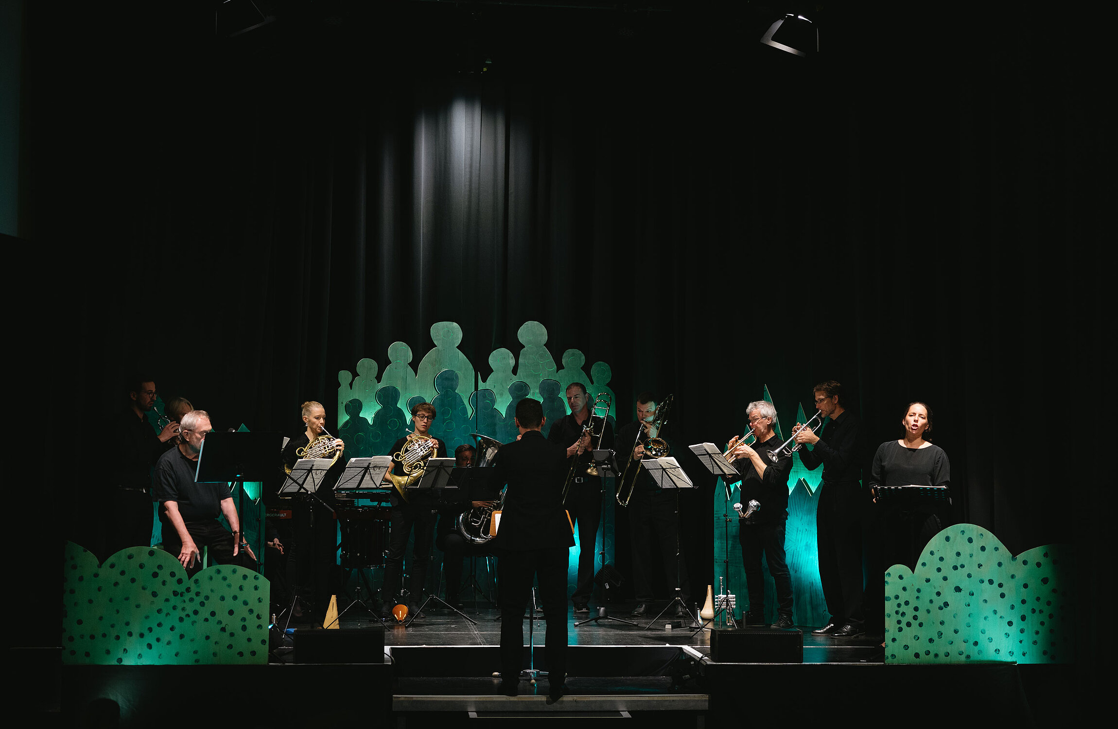 Blechblasensamble Brassersia spielt auf der Bühne mit einer Sopranistin und einem Sprecher vor grünem Bühnenbild.