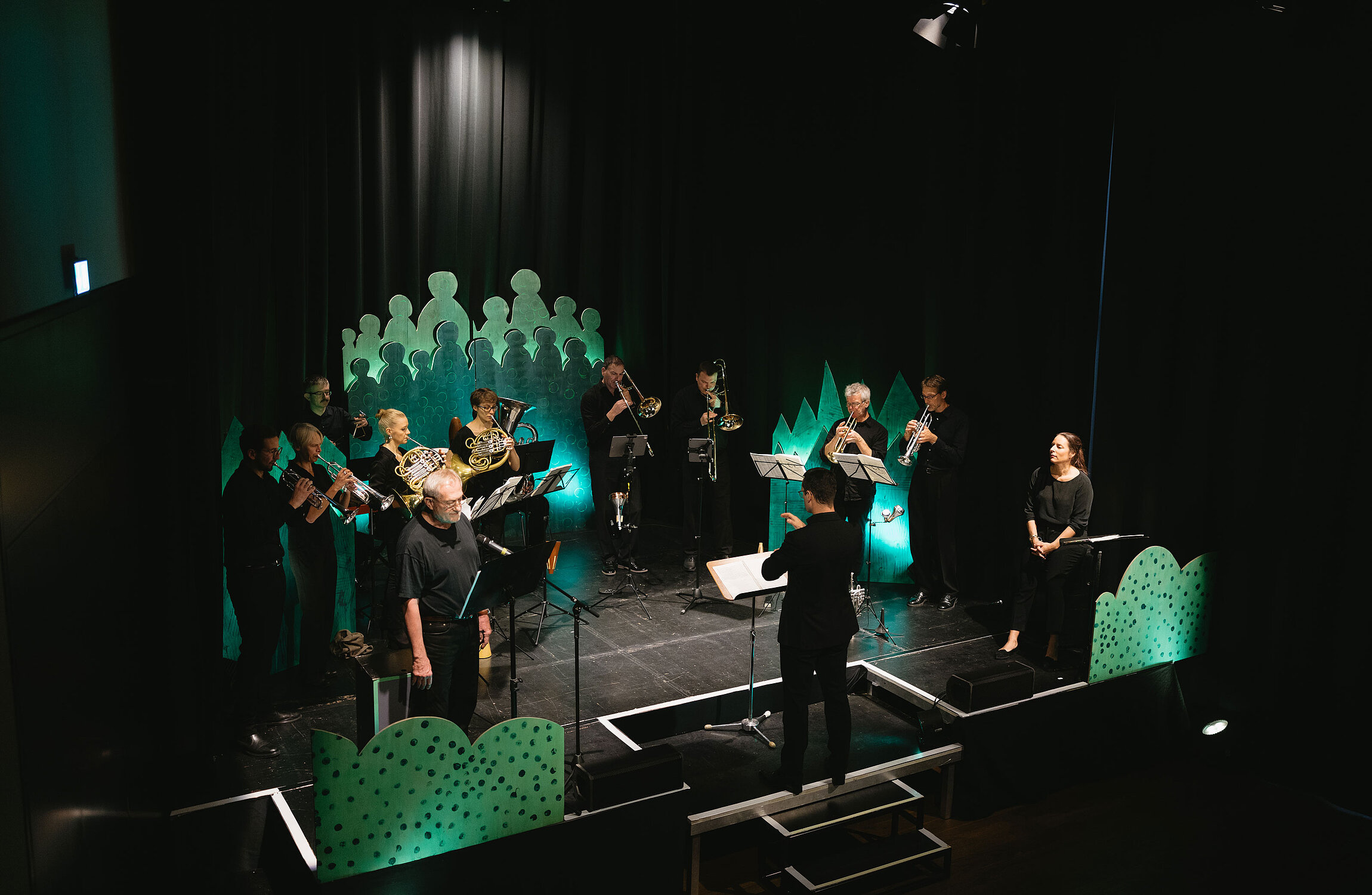 Blechblasensamble Brassersia spielt auf der Bühne mit einer Sopranistin und einem Sprecher vor grünem Bühnenbild.
