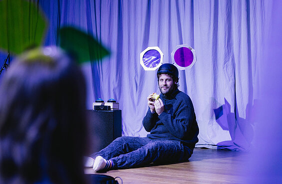 Schauspieler sitzt mit Helm mit Mauseohren und Apfel auf der Bühne vor blauem Bühnenvorhang.