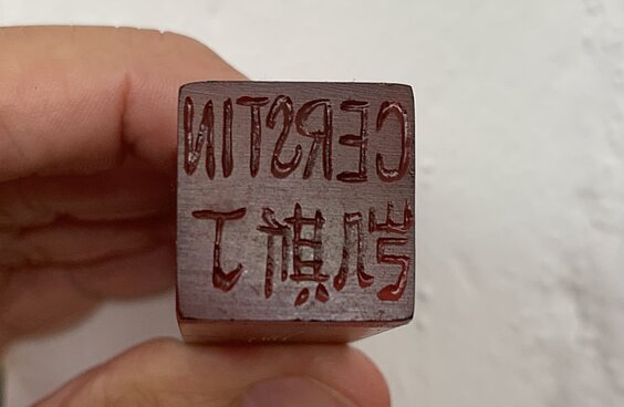 zwei Finger halten einen kleinen Stempel mit Initialien