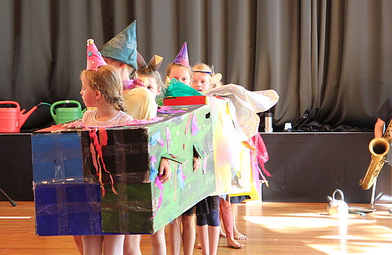 mehrere Kinder spielen in einem bunt bemalten Karton mit bunten Hüten begleitet von einem Saxophonisten und Gießkannen als Bühnenhintergrund