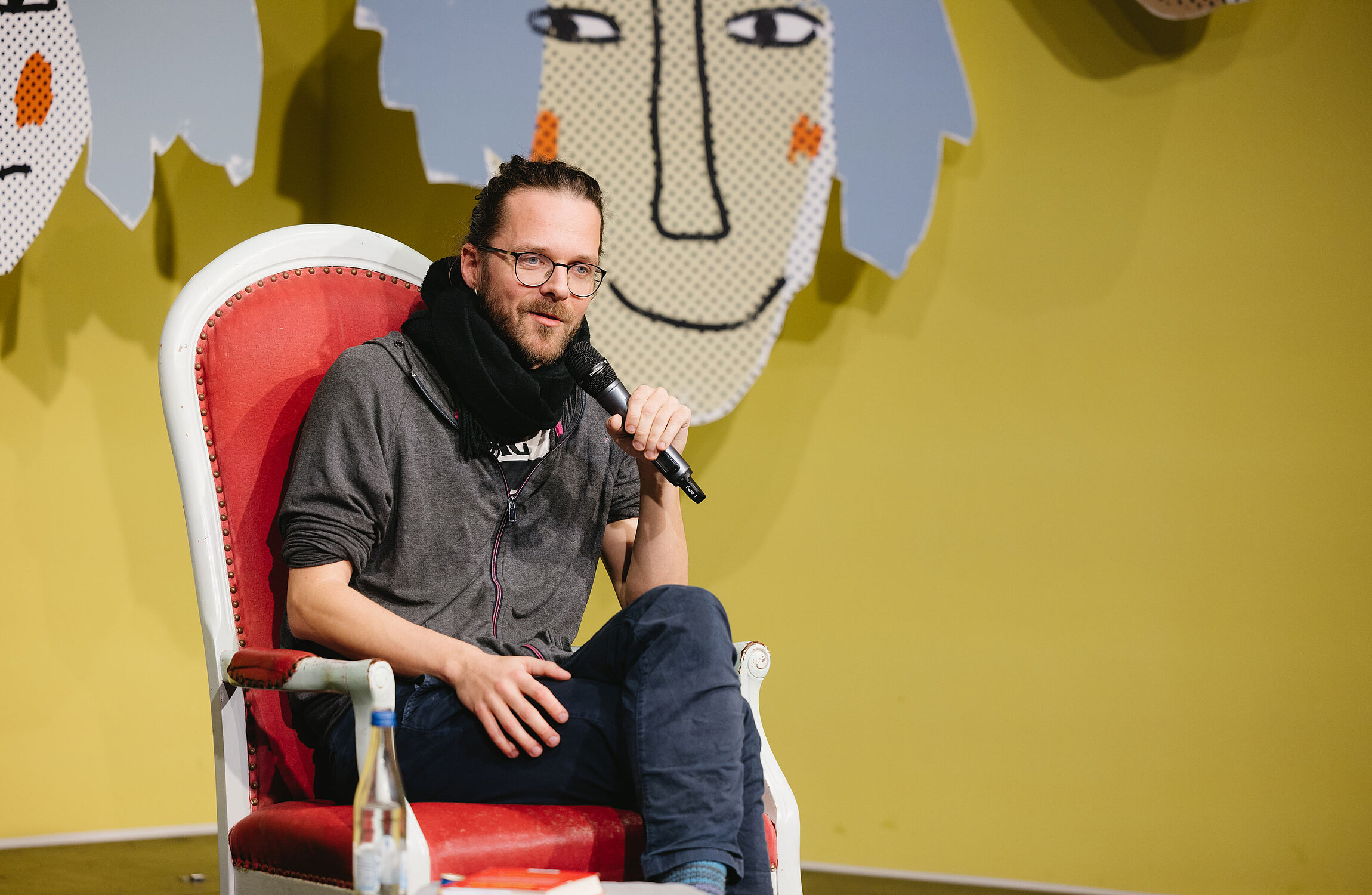 Der Autor Arne Semsrott hält ein Mikrofon und sitzt auf einem roten Sessel vor einer gelben Wand mit illustrierten Gesichtern.